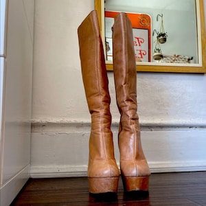 May include: A pair of knee-high tan leather boots featuring a platform sole and a block heel. The boots have a smooth finish, a rounded toe, and a vertical seam running down the center of the shaft. A side zipper is visible on the inner edge.