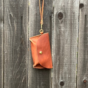 May include: A small, brown leather pouch with a curved flap and a brass button closure. It has a matching leather wrist strap attached with a gold-colored clasp. The pouch is hanging against a weathered wooden fence.