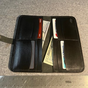 May include: Black leather wallet with multiple card slots and a billfold. The wallet is open and shows a credit card, a debit card, and a $20 bill. The wallet measures approximately 8 inches in length.