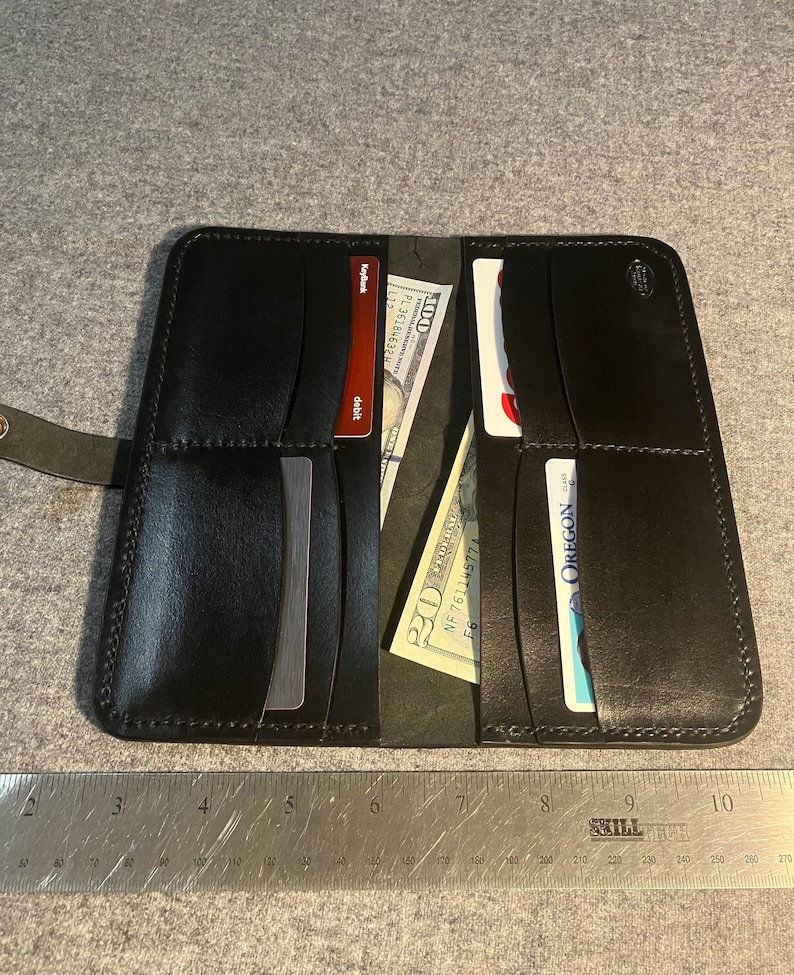 May include: Black leather wallet with multiple card slots and a billfold. The wallet is open and shows a credit card, a debit card, and a $20 bill. The wallet measures approximately 8 inches in length.