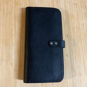 May include: A black leather wallet with a flap closure and a silver snap button. The wallet has a textured surface and a stitched edge.