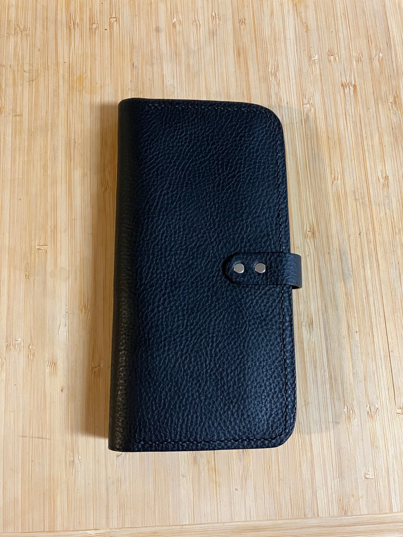May include: A black leather wallet with a flap closure and a silver snap button. The wallet has a textured surface and a stitched edge.