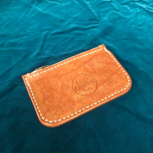 May include: A brown leather cardholder with a zipper closure and white stitching. The cardholder has a circular logo that reads "Grey Leather Co." The leather has a distressed appearance.
