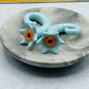 May include: Two light blue ceramic ear gauges with a star-shaped design and an orange center. The gauges are on a gray and white marble surface.