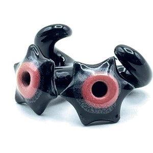 May include: Two black ceramic star-shaped beads with a red and white center hole. The beads have a glossy finish.
