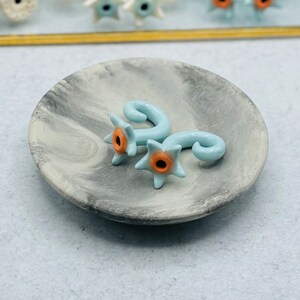 May include: Two light blue ceramic ear gauges with a star-shaped design and an orange center. The gauges are on a gray marble dish.