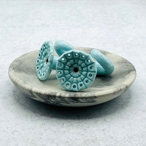May include: Three ceramic hooks in a light blue color with a detailed design. The hooks are sitting on a gray and white marble dish.