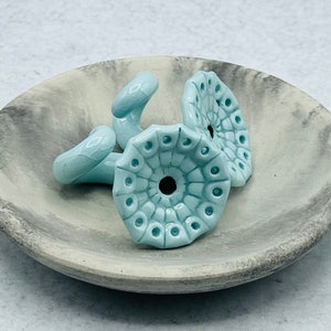 May include: A collection of light blue ceramic beads with a floral design, resting in a gray and white speckled ceramic bowl. The beads have a textured surface and a central hole.