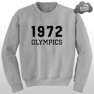 May include: A light grey crewneck sweatshirt featuring the black text 1972 OLYMPICS centred on the chest. A small grey cloud-shaped logo with the text the KING CONCEPT is positioned in the upper right corner of the garment.