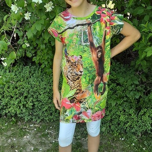 Könnte beinhalten: Ein Mädchen trägt ein grünes, weißes und rosa Kleid mit einem Dschungelprint, auf dem ein Orang-Utan und ein Leopard zu sehen sind. Das Kleid hat kurze Ärmel und einen runden Ausschnitt. Sie trägt eine weiße Leggings und blaue Sandalen.