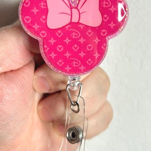 May include: Pink Minnie Mouse-shaped badge holder with a pink bow and a white and pink patterned design. The holder is attached to a clear plastic retractable badge reel.