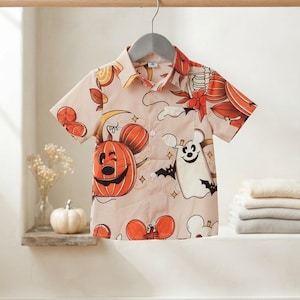 Puede incluir: Una camisa de manga corta con botones en color beige claro con un estampado repetitivo de temática de Halloween. El diseño incluye calabazas naranjas con forma de orejas de ratón, fantasmas blancos, pequeños murciélagos negros y hojas.