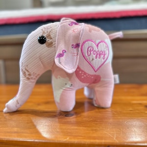 Op de afbeelding: Roze stoffen olifant knuffel met een hartvormig patch waarop "Poppy" staat. De olifant heeft een roze en bruine gepatenteerde stof met afbeeldingen van flamingo's, ijsberen en andere dieren.