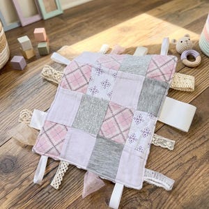 May include: A square sensory tag blanket featuring a patchwork design of light pink, grey, and white fabric squares. The fabric patterns include argyle, small floral prints, and solid colors. Multiple fabric ribbons and lace trims extend from the edges.
