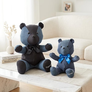 May include: Two denim teddy bears with black and blue accents sit on a table. The larger bear has a black torso and bow tie, while the smaller bear has a blue bow. Both bears have the name "Torrey" embroidered on their feet.