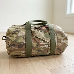 May include: A cylindrical duffel bag featuring a multi-colored camouflage pattern in shades of green, brown, and tan. The bag includes olive green nylon webbing handles that wrap around the body and a small red rectangular tag on the top.