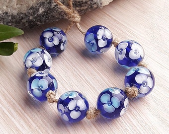 Puede incluir: Un collar de cuentas de vidrio azul y blanco con flores blancas en cada cuenta. El collar está encordado en un cordón de cuerda natural.