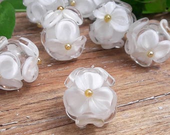 May include: White glass beads with a flower design and gold accents. The beads are arranged on a wooden surface.