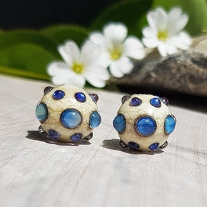 May include: Two spherical beads featuring a cream-colored base with a fine crackle texture. Each bead is decorated with multiple circular inlays in varying shades of blue, ranging from light sky blue to deep navy, set within dark metal rims.