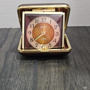 May include: A vintage travel alarm clock in a brown leatherette case, open to reveal a square clock face. The clock face is brown with gold numerals and hands, and the word "Linden" is printed on the face.