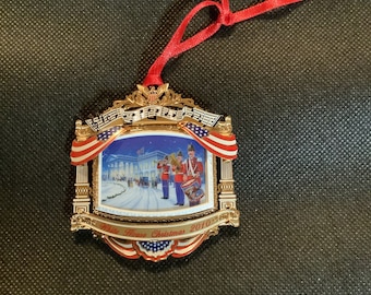 May include: A gold-colored Christmas ornament with a red ribbon. The ornament features a framed image of the White House with a marching band. The frame is decorated with red, white, and blue accents and the text "White House Christmas 2010".