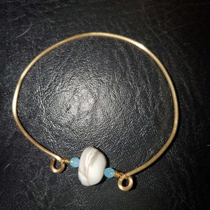 May include: A gold-colored bangle bracelet with a white and gray stone bead and two small blue beads. The bracelet is on a black surface.