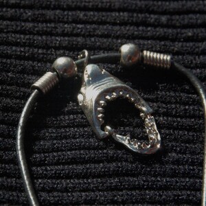 May include: A silver shark tooth pendant with a detailed open mouth, attached to a black cord necklace. The pendant is accented with silver beads and coiled metal elements. The necklace is displayed against a textured black background.