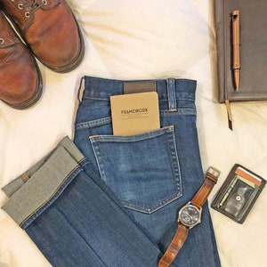 May include: A pair of blue jeans with a brown leather watch and a small brown notebook with the word "FRAMEWORK" on the cover. The jeans are folded and the watch is laying on top of them.