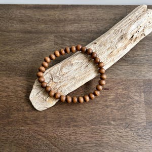 May include: A brown wooden bead bracelet resting on a piece of driftwood. The bracelet is made of small, round beads and is displayed on a dark brown wooden surface. The driftwood is light brown and has a rough texture.