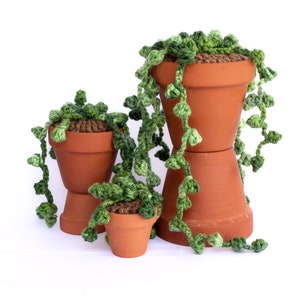 May include: Three crocheted succulent plants in terracotta pots. The plants are green and have long trailing stems. The pots are stacked on top of each other.