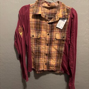 May include: A maroon and plaid long-sleeve shirt. The shirt features a plaid pattern in shades of brown and yellow on the front and collar, with maroon sleeves. The left sleeve has a gold emblem. The shirt is hanging on a metal hanger.