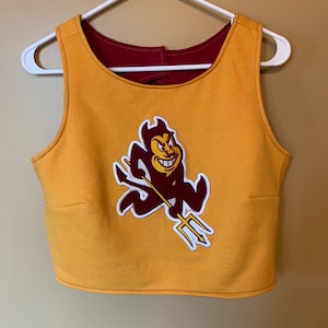 Könnte beinhalten: Ein wendbares Crop-Top in Gold mit bordeauxroter Innenseite. Auf der Vorderseite befindet sich ein Cartoon-Teufel-Maskottchen in Burgunderrot und Weiß, das einen Dreizack hält. Das Oberteil hängt an einem weißen Kleiderbügel.