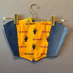 Könnte beinhalten: Ein Jeans- und gelbes Korsett-Top mit dem Text "Go Devils" und einem Handsymbol. Das Top hängt an einem goldenen Kleiderbügel. Der gelbe Stoff befindet sich in der Mitte, mit Jeans auf beiden Seiten.