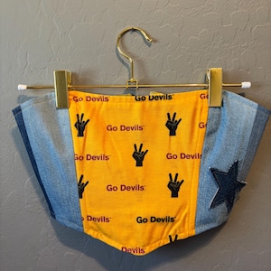 Könnte beinhalten: Ein Bandeau-Top aus Jeansstoff und Gelb mit dem Text "Go Devils" und einer Peace-Zeichen-Grafik. Das Top hat einen dunkelblauen Sternaufnäher an der Seite und hängt an einem goldenen Kleiderbügel.