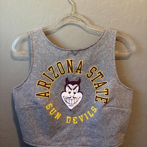 Könnte beinhalten: Graues Crop-Top mit den Worten "ARIZONA STATE SUN DEVILS" in gold- und kastanienbraunen Buchstaben. Das Oberteil zeigt ein Cartoon-Teufel-Maskottchen in der Mitte. Das Oberteil hängt an einem durchsichtigen Kunststoffbügel.