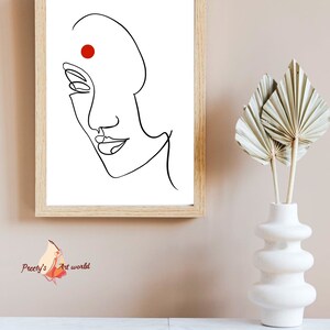 May include: A black and white line drawing of a woman's face with a red dot on her forehead. The drawing is framed in a light wood frame and is hanging on a light pink wall.