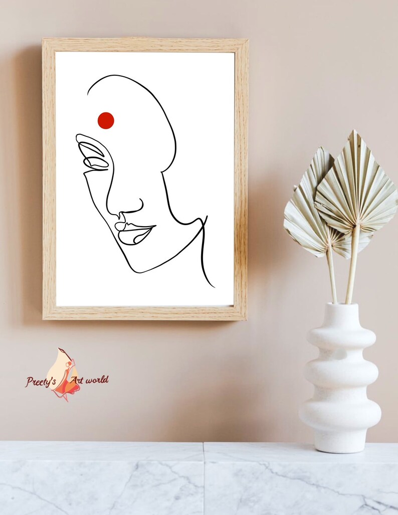 May include: A black and white line drawing of a woman's face with a red dot on her forehead. The drawing is framed in a light wood frame and is hanging on a light pink wall.