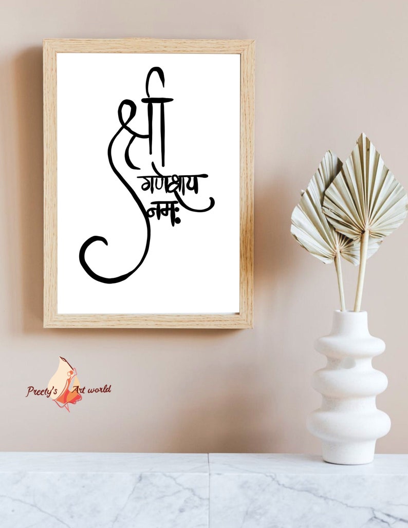 May include: A framed print with a black and white calligraphy design that says "Shri Ganeshay Namah". The print is hanging on a light brown wall with a white vase and dried palm leaves in the foreground.