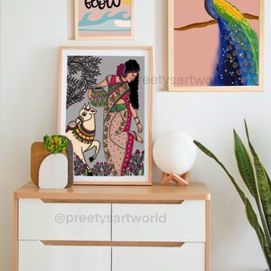 May include: A framed illustration of a woman in a colorful sari holding a pot, with a white cow beside her. The illustration is set against a gray background with green trees and flowers. The frame is wooden and has a white mat.