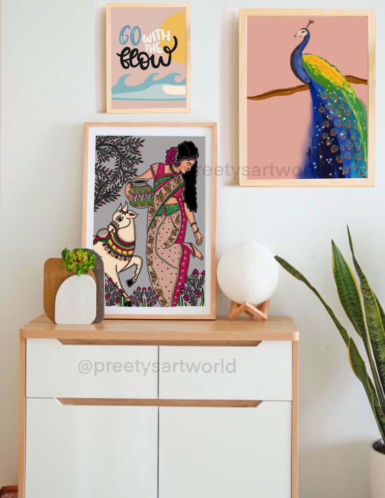 May include: A framed illustration of a woman in a colorful sari holding a pot, with a white cow beside her. The illustration is set against a gray background with green trees and flowers. The frame is wooden and has a white mat.