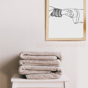 May include: A black and white line drawing of two hands holding each other. The hand on the left is wearing a stack of bangles. The drawing is framed in a light wood frame and hanging on a white wall.