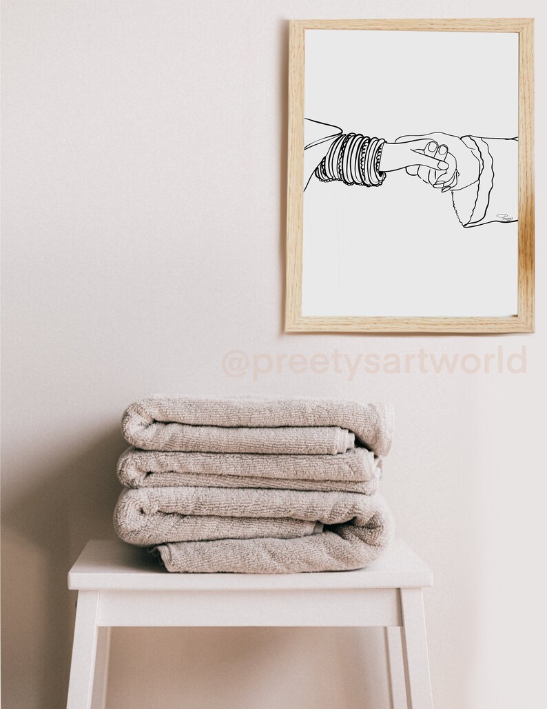 May include: A black and white line drawing of two hands holding each other. The hand on the left is wearing a stack of bangles. The drawing is framed in a light wood frame and hanging on a white wall.