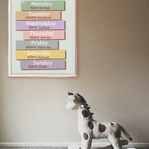 Puede incluir: Un gr&aacute;fico colorido con los d&iacute;as de la semana en ingl&eacute;s y punjabi. El gr&aacute;fico est&aacute; enmarcado y colgado en una pared. Un caballo de balanc&iacute;n de peluche con forma de jirafa est&aacute; en primer plano.