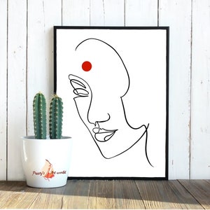 May include: A black and white line drawing of a woman's face with a red dot on her forehead. The drawing is framed in a black frame and is hanging on a white wall.
