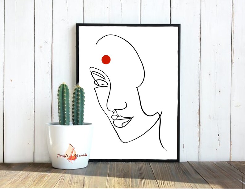 May include: A black and white line drawing of a woman's face with a red dot on her forehead. The drawing is framed in a black frame and is hanging on a white wall.