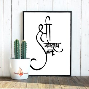 May include: Black and white calligraphy print with the text "Shree Ganeshay Namah" in a decorative font. The print is framed in a black frame and is displayed on a white wooden wall.
