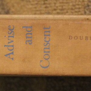 Op de afbeelding: Een bruine boekrug met de titel "Advise and Consent" van Allen Drury en de uitgever Doubleday.