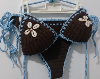 May include: A handmade brown crochet bikini set with light blue trim and ties. The bikini top features seashell embellishments. The set is displayed on a white hanger against a white background. A unique, beach-themed swimwear item.