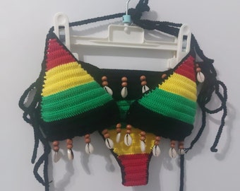 May include: A handmade crochet bikini set in the colors of red, yellow, and green, with black trim. The bikini top and bottom are adorned with wooden beads and small seashells. The set is hanging on a white hanger against a white background.