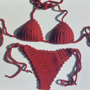 May include: A handmade red crochet bikini set. The top features triangle cups with adjustable ties, and the bottoms have a low-rise design with tie sides. The crochet work has a textured, ribbed pattern.